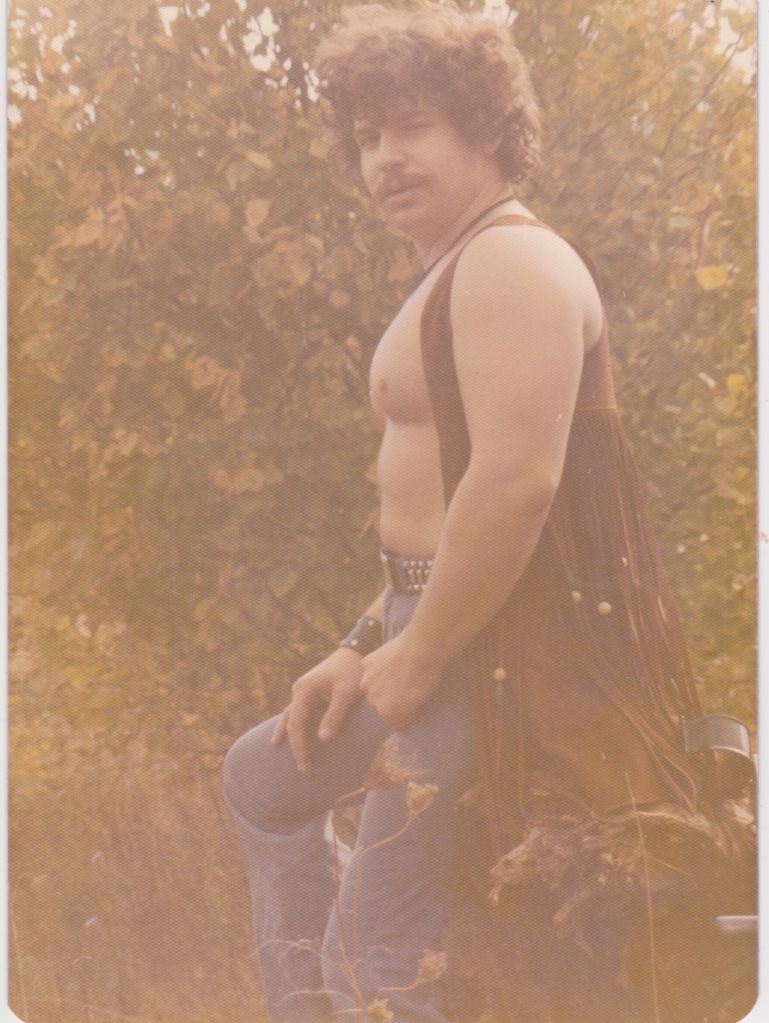 Bare chested picture of me sitting on a tree stump adorned with a Janis Joplin type leather vest, lots of leather tassels with beads,  